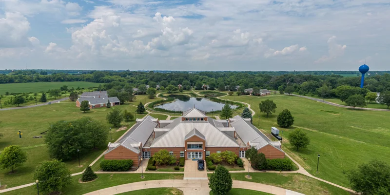 Aerial view of a peaceful, expansive campus with lush greenery, walking paths, and a serene pond, offering a tranquil environment.