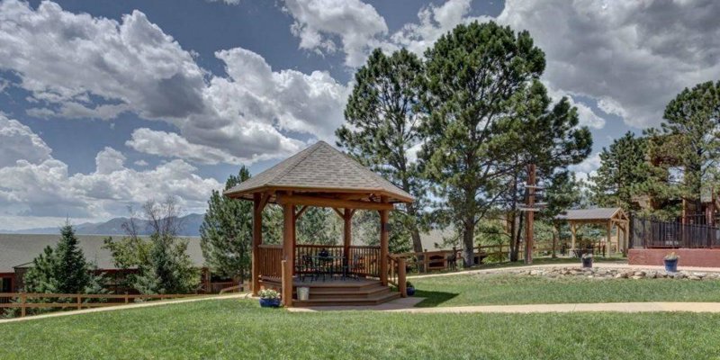 Charming gazebo surrounded by lush greenery and towering trees, set against a backdrop of serene mountains and a bright, open sky.