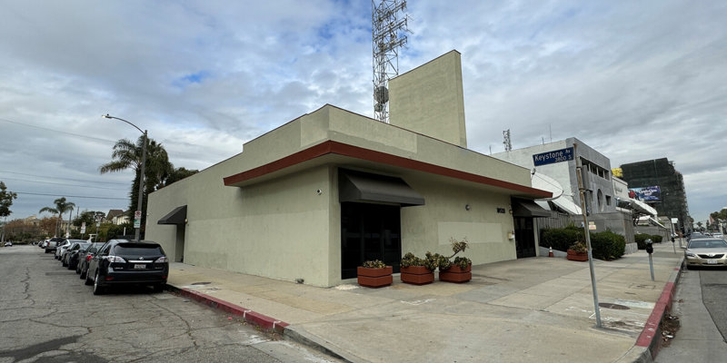 A modern, single-story building with a beige exterior and red trim, located on a corner lot, with several cars parked along the street.