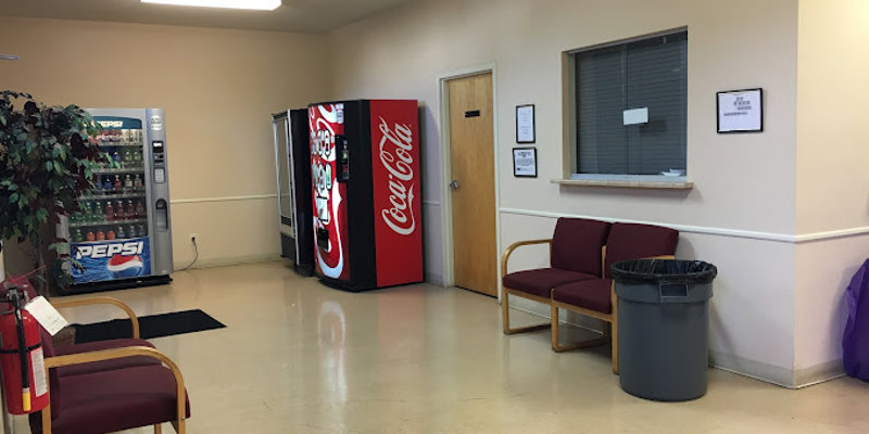 A casual waiting area with vending machines, red cushioned chairs, and a trash bin. The space is bright and clean, offering refreshments for visitors.