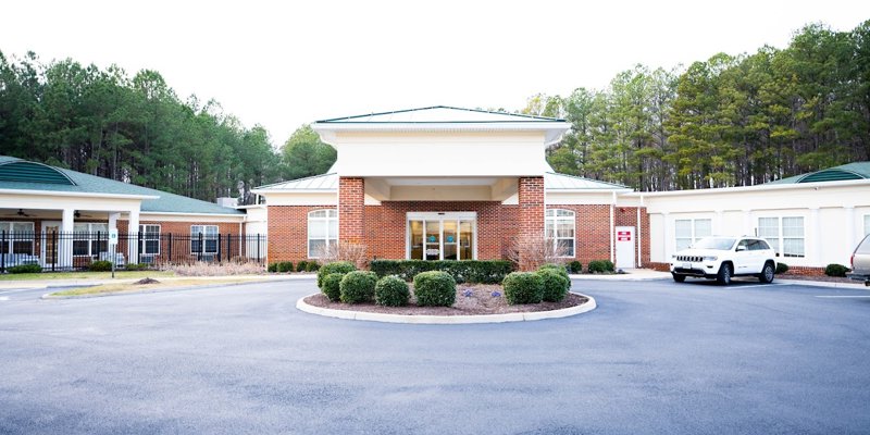 Inviting entrance to a brick facility with landscaped greenery, a circular driveway, and adjacent buildings surrounded by lush trees.