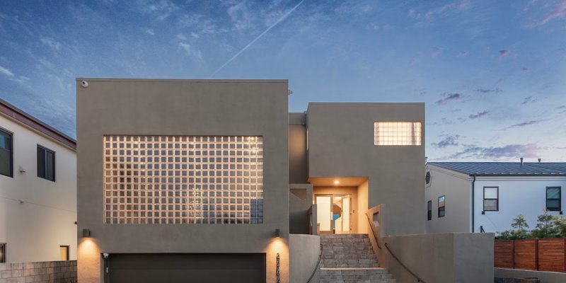 A modern two-story home with sleek lines, illuminated by warm exterior lights, features a large glass block window and an inviting staircase.