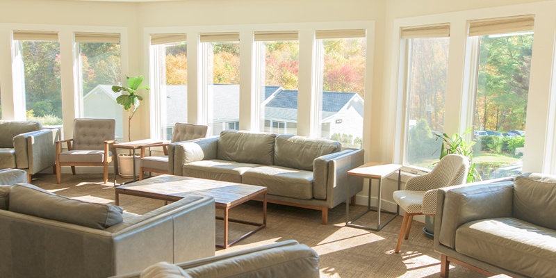 A bright and inviting lounge area featuring cozy gray sofas, natural wood tables, large windows, and scenic outdoor views bathed in sunlight.