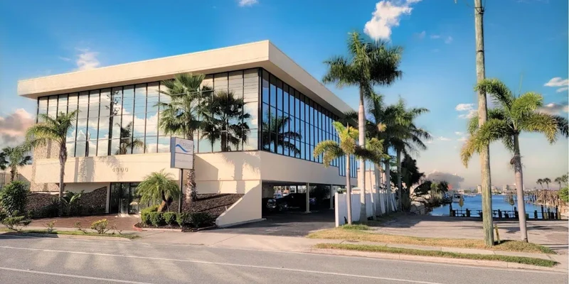  A sleek, modern two-story building with large glass windows, surrounded by palm trees, located near a peaceful waterfront under a clear sky.