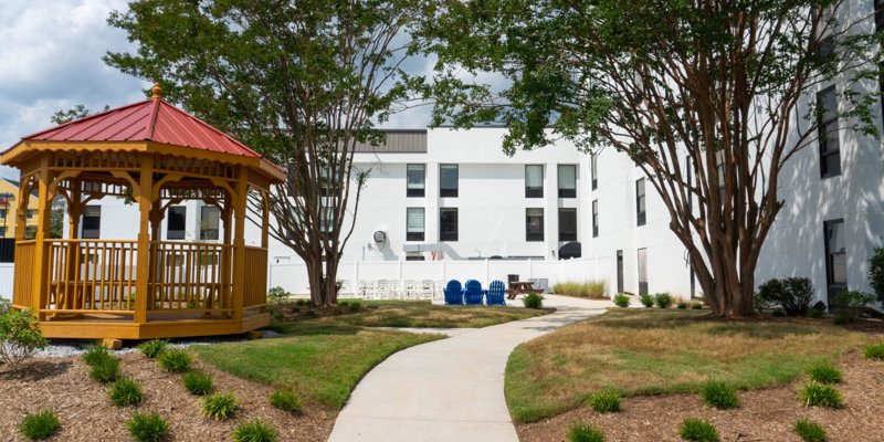 A peaceful outdoor area featuring a charming gazebo, shaded trees, and a pathway leading to cozy seating, perfect for relaxation and reflection.