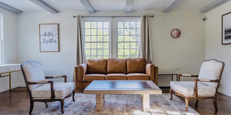 Cozy seating area with a stylish brown sofa, classic armchairs, and rustic wood-beamed ceiling, bathed in natural light from large windows.