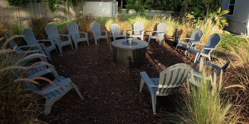  A tranquil outdoor gathering space with a circle of Adirondack chairs around a fire pit, surrounded by lush greenery, perfect for group relaxation.