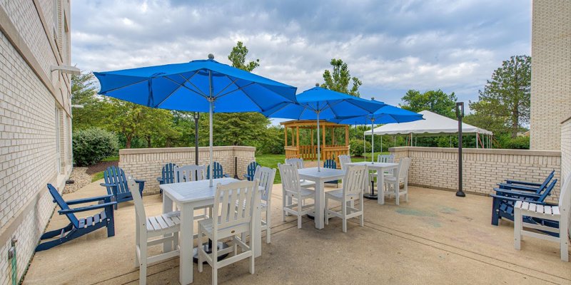 A charming outdoor patio with white tables and chairs, shaded by vibrant blue umbrellas, surrounded by greenery for a relaxing atmosphere.