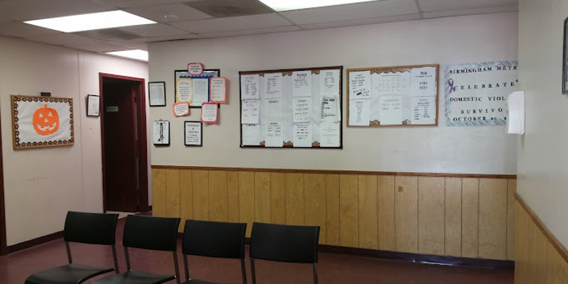 A simple waiting area with black chairs, wood-paneled walls, and bulletin boards displaying various notices and information. The space is well-organized and welcoming.