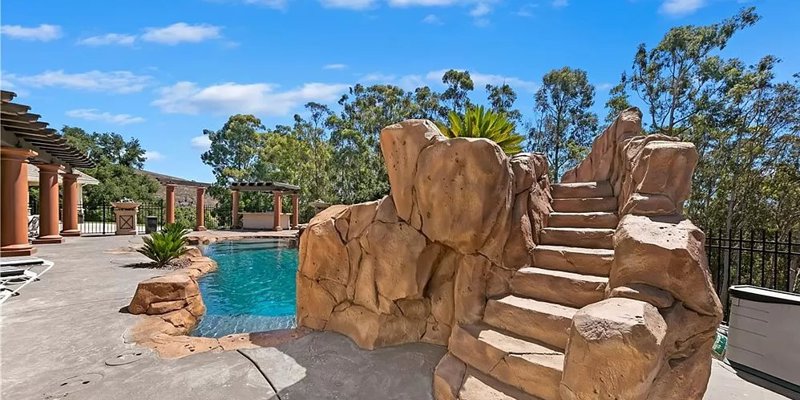 A beautiful pool area with natural rock features and stone steps leading to a water slide, set amidst lush greenery for a resort-like feel.