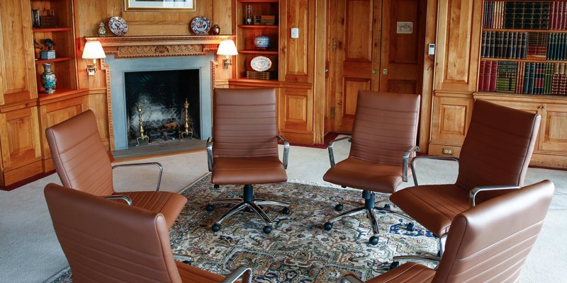 Warm and elegant meeting room featuring leather chairs arranged in a circle, a cozy fireplace, wood-paneled walls, and built-in bookshelves.