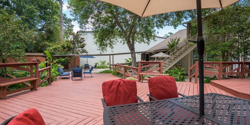 A peaceful outdoor deck with red cushioned chairs, an umbrella for shade, and cozy seating areas, surrounded by lush greenery and trees.