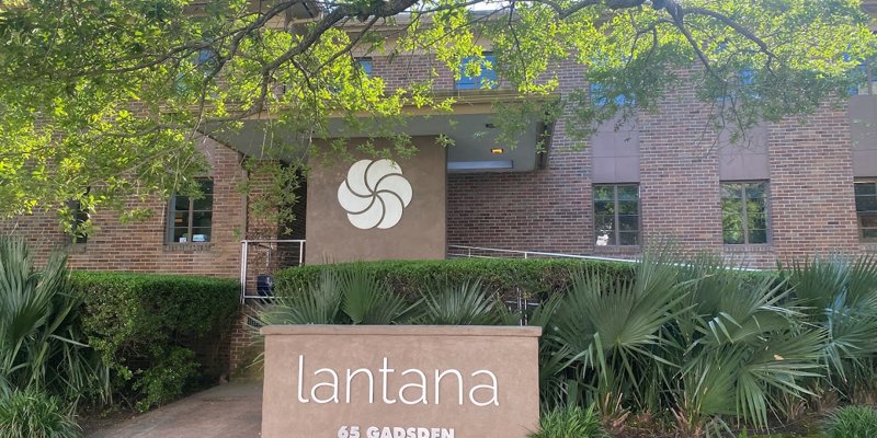 Welcoming entrance to Lantana, surrounded by lush greenery and shaded by trees, with a modern sign and a well-maintained brick building.