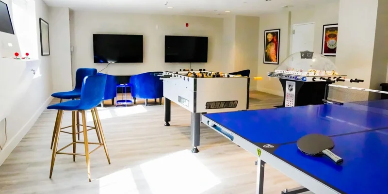 A bright and welcoming game room featuring foosball, ping-pong, and arcade games, with comfortable blue seating and entertainment screens.