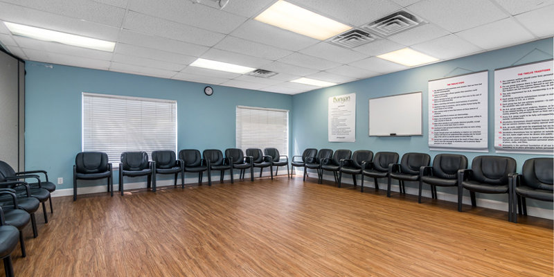 A large group room with black chairs arranged in a circle on a wood floor. The walls are light blue, featuring inspirational posters and a whiteboard.