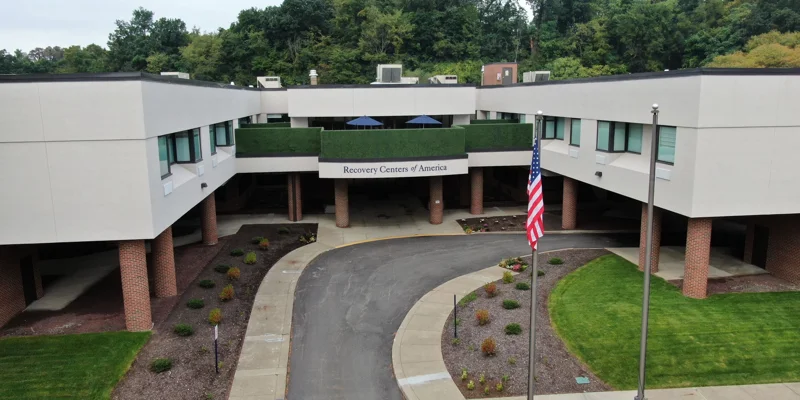 A modern recovery center with a circular driveway, clean landscaping, and an American flag, surrounded by lush greenery in a peaceful setting.