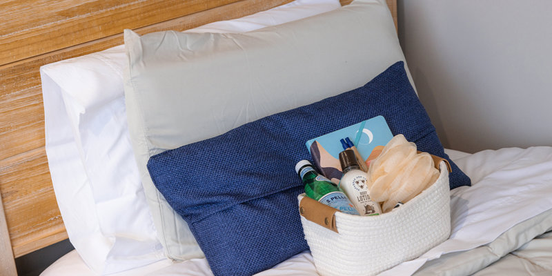  A cozy bed with soft pillows and a welcoming gift basket, including lotion, water, and a loofah, offering comfort and relaxation.