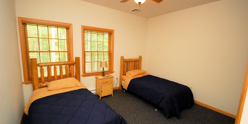 A cozy bedroom featuring two wooden twin beds with navy bedding, warm lighting, and large windows that invite natural light.