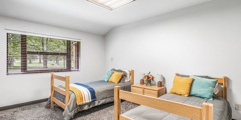 Bright and cozy shared bedroom with soft lighting, neatly made beds, colorful pillows, and a large window overlooking peaceful greenery.