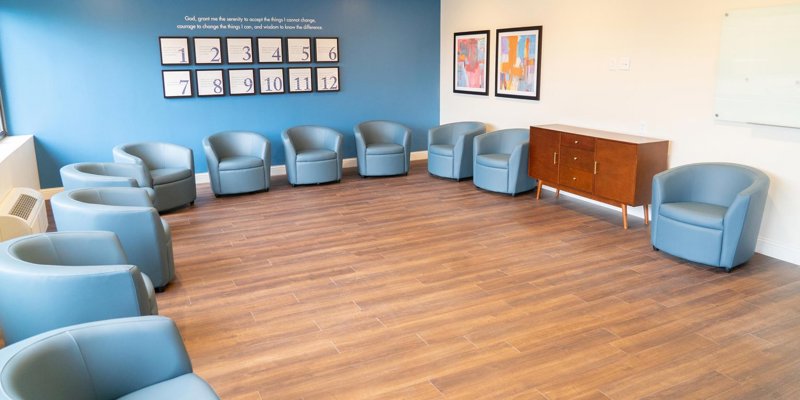 A bright group therapy room with comfortable blue chairs arranged in a circle, wood flooring, and motivational decor on a calming blue accent wall.