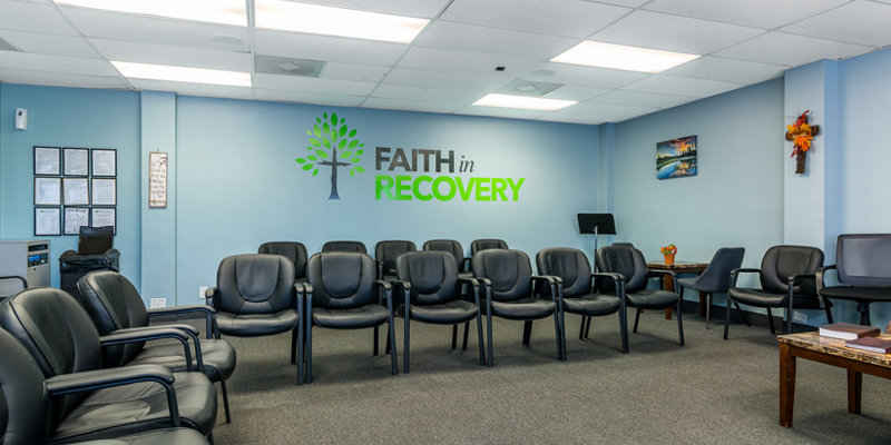 A spacious and comfortable meeting room with rows of black chairs and a bright "Faith in Recovery" sign on the wall.