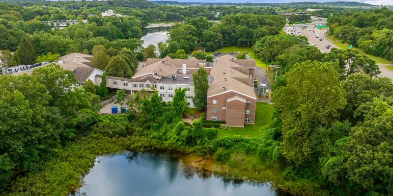 Aerial view of a tranquil recovery center surrounded by lush greenery, a serene pond, and nearby highway access, blending nature with convenience.