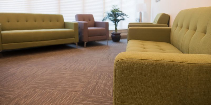 A comfortable seating area with green and beige sofas, soft carpeting, and natural light coming through large windows, creating a relaxing atmosphere.