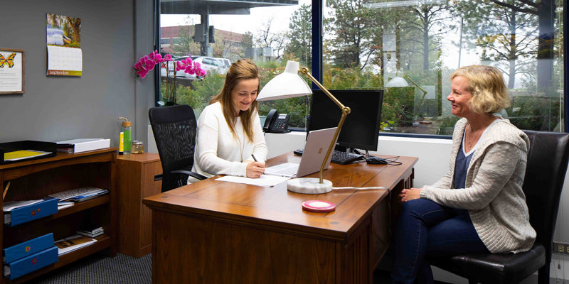 A cheerful therapist writes notes while a client sits comfortably, engaged in conversation, within a bright and welcoming office space.