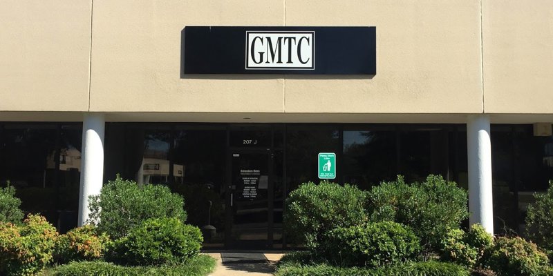 A simple, welcoming building with a clean exterior, a "GMTC" sign above the entrance, and neatly trimmed bushes lining the pathway.