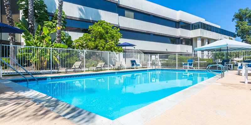 A pristine swimming pool surrounded by lush greenery and shaded seating areas, perfect for relaxation and rejuvenation under the bright blue sky.