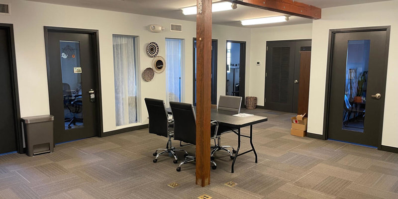  A modern office space with private meeting rooms, a central table with chairs, exposed wooden beams, and clean, neutral decor, offering a professional environment.