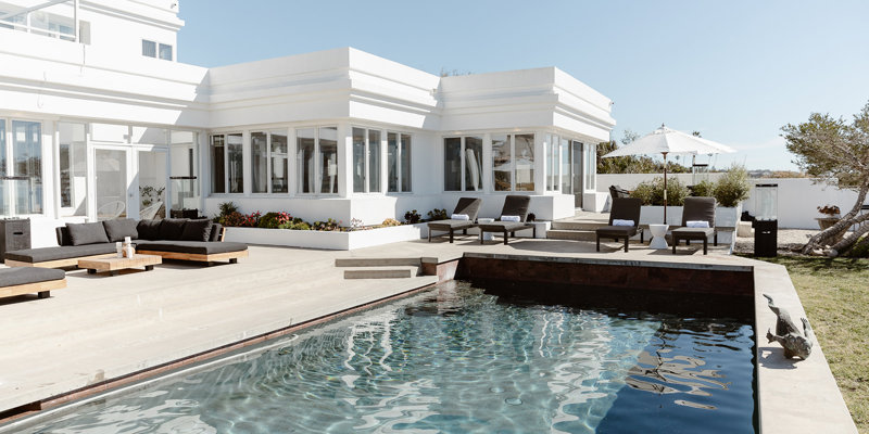  Elegant poolside patio featuring modern lounge chairs, shaded umbrellas, and sleek white architecture, offering a serene oceanfront view.