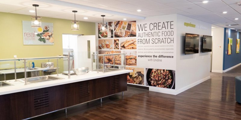 A bright and modern dining area with a clean food counter, vibrant signage promoting fresh, authentic meals made from scratch, and wood flooring.