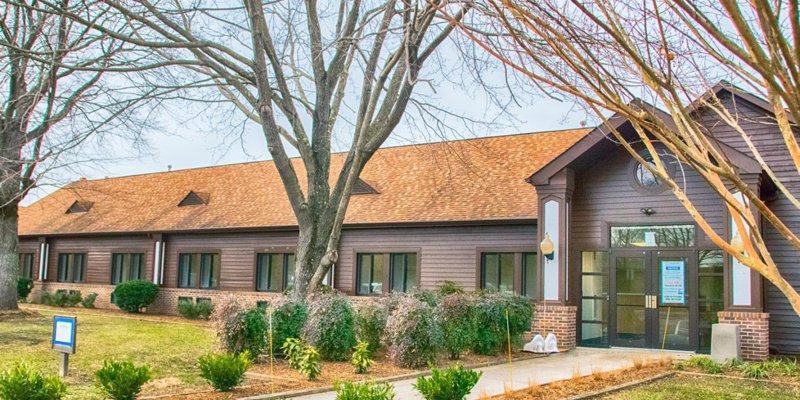 A charming building with a sloped roof and large windows, surrounded by neatly trimmed bushes and trees. The peaceful setting enhances its welcoming atmosphere.