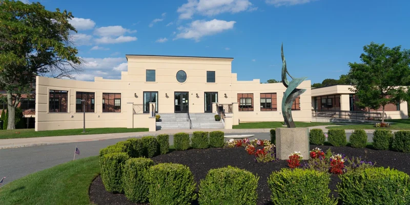 A beautifully landscaped building entrance with trimmed hedges, vibrant flowers, and a modern sculpture stands under a bright blue sky.