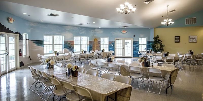 A spacious dining area with round and rectangular tables, decorated with flowers, featuring a serene beach mural and bright natural light.