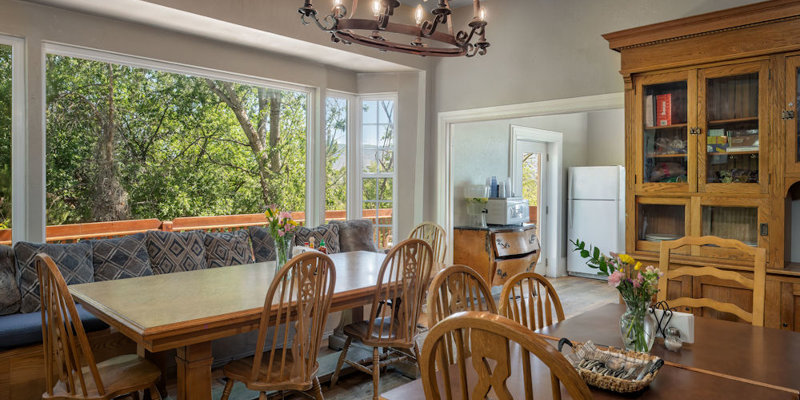 A cozy dining area with wooden tables and chairs, large windows showcasing a lush green view, and a warm chandelier above. A homey atmosphere.