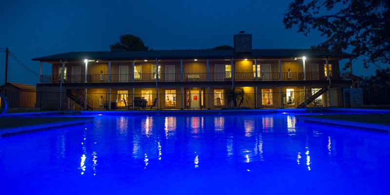 A beautifully illuminated pool at night reflects the warm glow of a cozy two-story building, creating a peaceful and inviting ambiance.