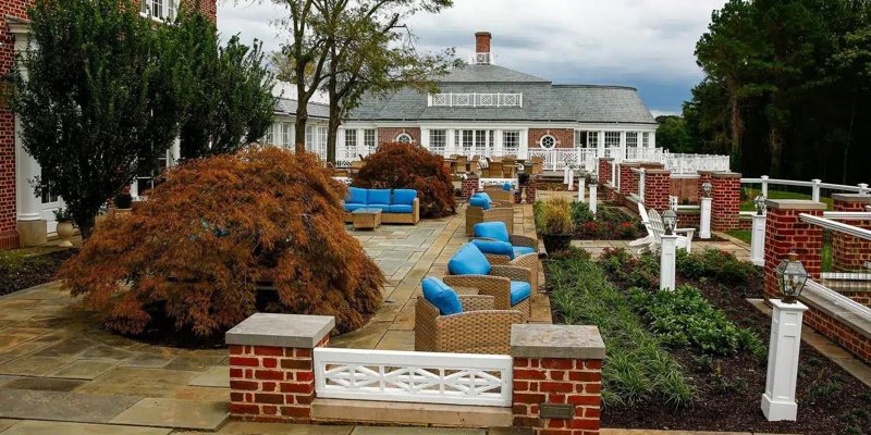 Elegant outdoor patio with cozy wicker seating featuring blue cushions, surrounded by manicured gardens, brick accents, and lush greenery.