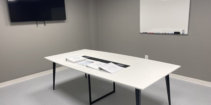  A clean, well-organized meeting room with a large white table, binders, a mounted TV, and a whiteboard, perfect for group discussions or presentations.