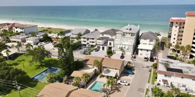 Aerial view of a serene beachfront neighborhood, featuring homes, lush green spaces, and clear ocean waters under a bright, blue sky.