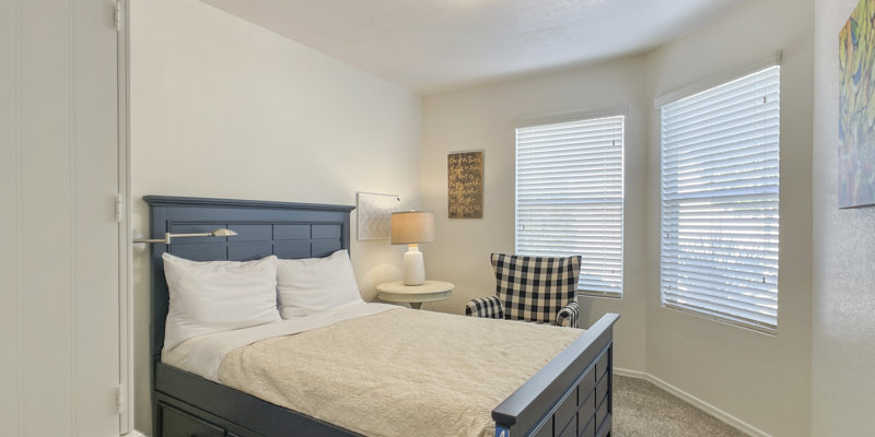 Cozy bedroom with soft natural light, featuring a comfortable bed with white linens, a checkered armchair, and a warm bedside lamp.