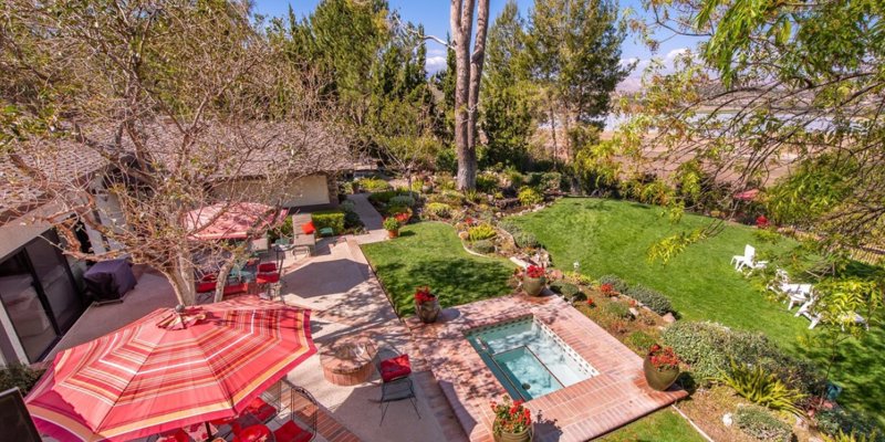  A beautifully landscaped backyard with a hot tub, lush green lawn, colorful flowers, and cozy seating areas under vibrant umbrellas.