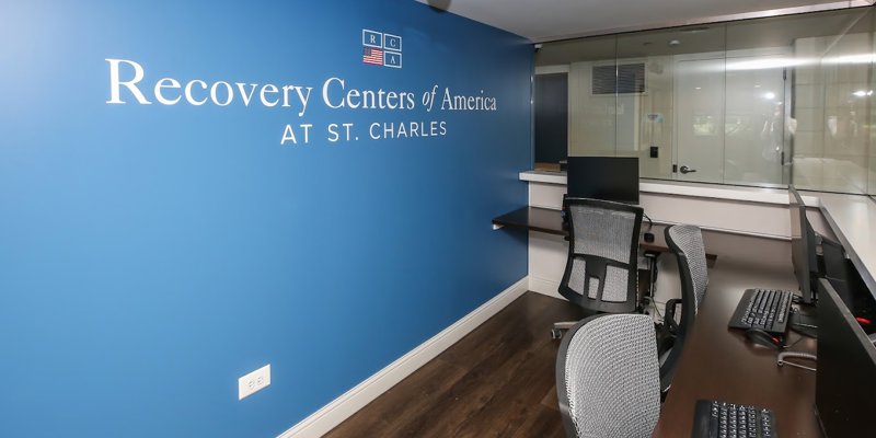 Modern workstations with ergonomic chairs and computers, set against a blue accent wall featuring the Recovery Centers of America logo.