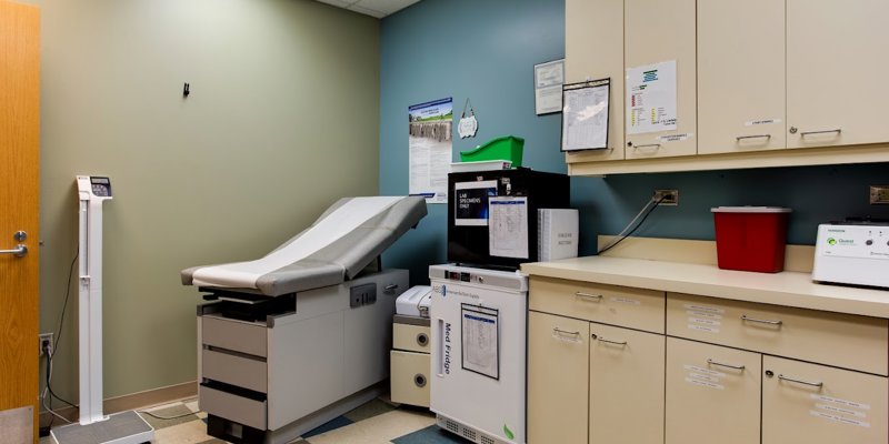 A clean and organized medical examination room featuring a padded exam table, cabinets, a scale, and essential equipment for patient care.