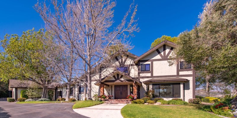 A picturesque two-story home with charming architectural details, surrounded by lush greenery and colorful flowers, under a clear blue sky.