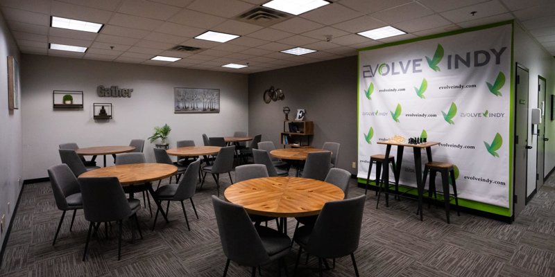 Modern gathering space with round wooden tables, comfortable gray chairs, and a branded backdrop, ideal for group activities and meetings.