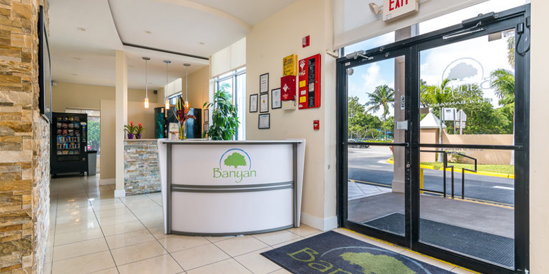 A bright and welcoming reception area with the Banyan logo on the desk and entrance mat. The space is modern, with stone accents, plants, and large windows letting in natural light.