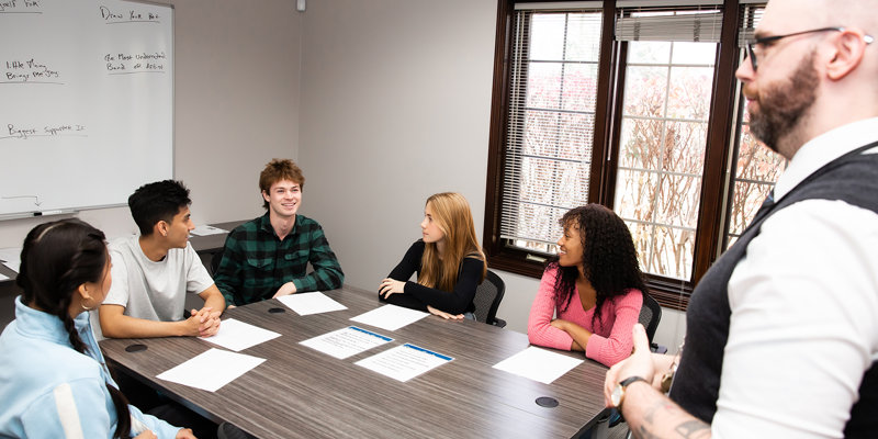 A group of young individuals sits around a table in discussion, guided by an instructor, creating a collaborative and engaging environment.