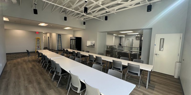 A bright and spacious dining area with long tables, comfortable seating, and modern lighting, offering a welcoming environment for meals or group gatherings.